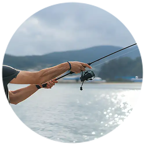 A fisherman casting a fishing rod over calm waters, wearing waterproof bracelets, with mountains and a cloudy sky in the background.