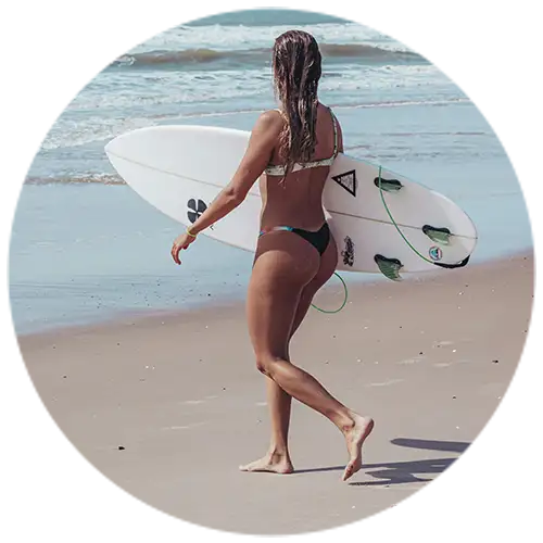 A female surfer walking along a beach with a surfboard, wearing a bracelet and bikini, showcasing waterproof jewelry with the ocean in the background.