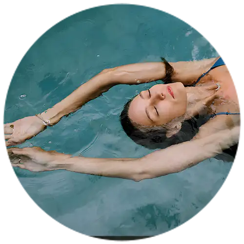 A woman floating on her back in a pool, wearing a bracelet and a necklace, with arms stretched out gracefully, surrounded by calm blue water.