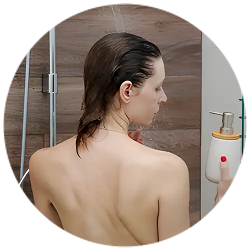 A woman with wet hair wearing a delicate necklace in a shower, holding a soap dispenser, set in a modern bathroom with wood-like tiles.