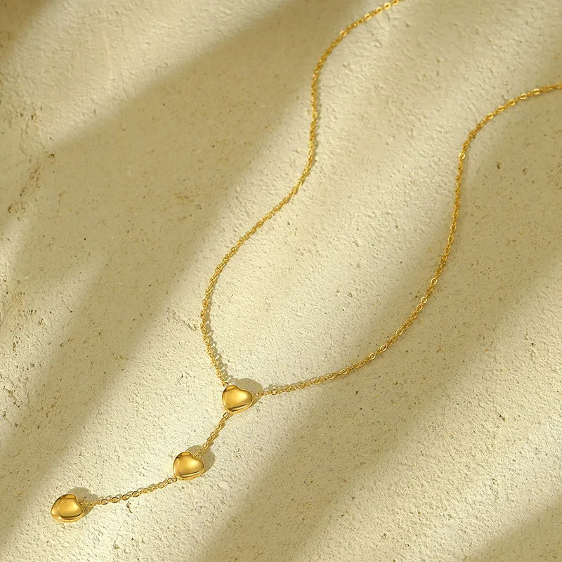 A gold heart drop necklace with three smooth heart charms, displayed on a textured beige surface under soft sunlight.