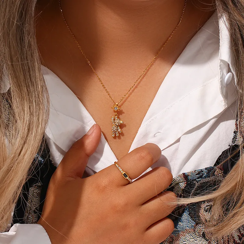 A person wearing a gold teddy bear pendant necklace with cubic zirconia and a matching star charm, paired with a simple gold ring, styled with a white shirt.