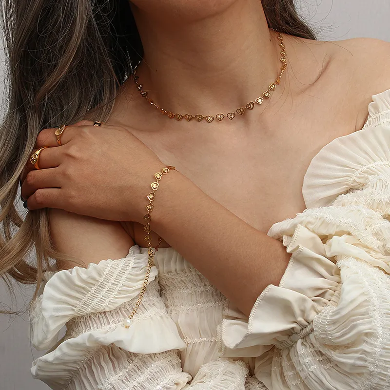 Model wearing an 18K gold-plated heart chain jewelry set, including a choker necklace, bracelet, and rings, styled with a romantic off-shoulder white dress.
