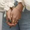 Close-up of hands adorned with an 18K gold-plated heart chain bracelet and statement rings, styled with a ruffled blouse and denim jeans.