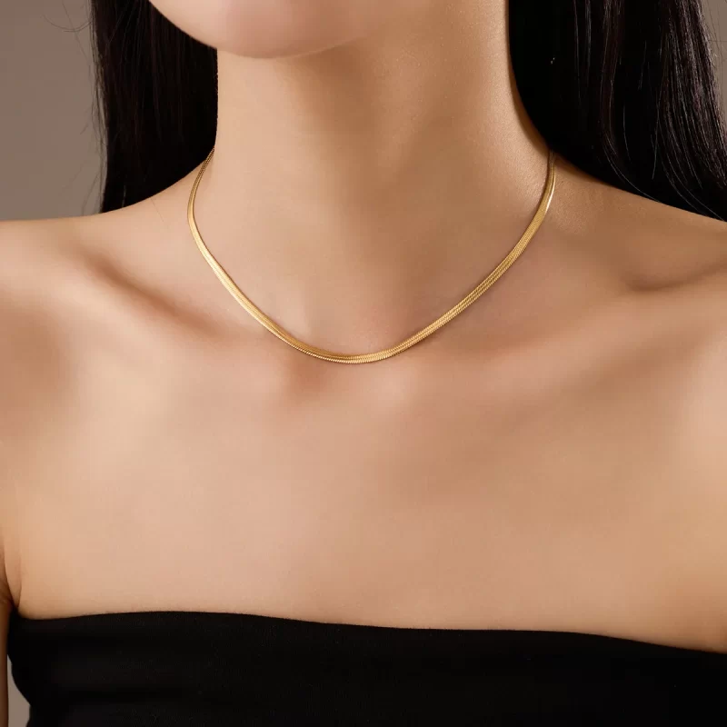 A gold herringbone chain necklace with a polished finish, elegantly worn on a model with a black strapless top.
