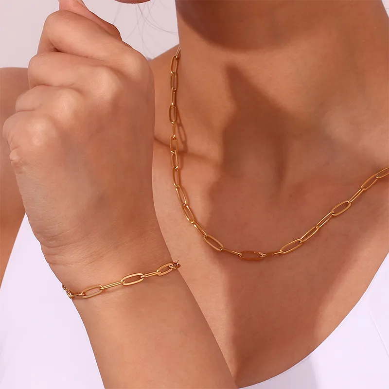 A woman wearing a gold paperclip chain necklace and matching bracelet, featuring elongated oval links, against a soft pink background.