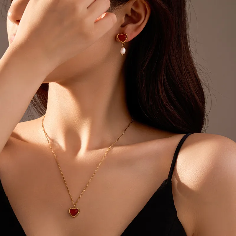 A woman wearing a gold-plated red agate heart necklace and matching pearl drop earrings, styled with a black outfit against a soft-lit background.