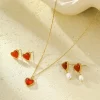 A set of heart-shaped red agate jewelry, including a gold-plated necklace, stud earrings, and pearl drop earrings, displayed on a textured neutral background.