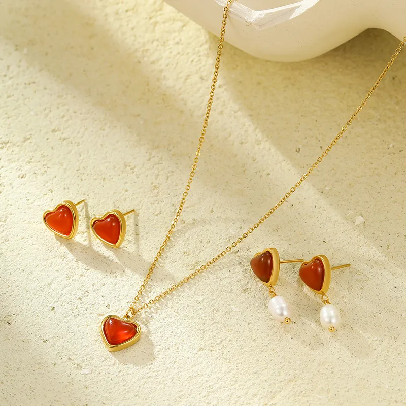 A set of heart-shaped red agate jewelry, including a gold-plated necklace, stud earrings, and pearl drop earrings, displayed on a textured neutral background.