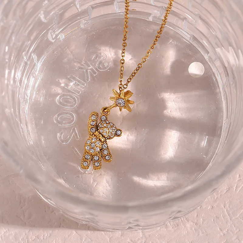 A gold teddy bear pendant necklace with cubic zirconia stones and a star charm inside a clear glass container.