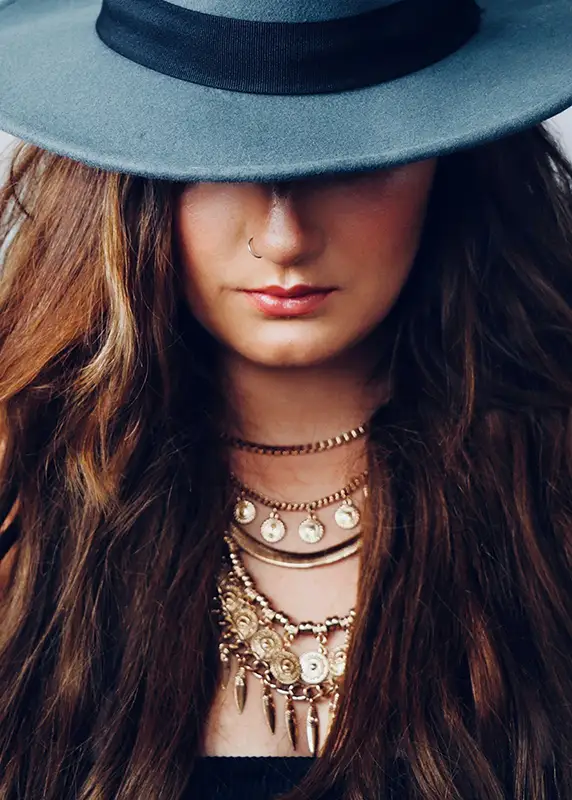 A woman with long wavy hair wearing a wide-brim hat and layered gold necklaces with coins and pendants, creating a boho-chic look.