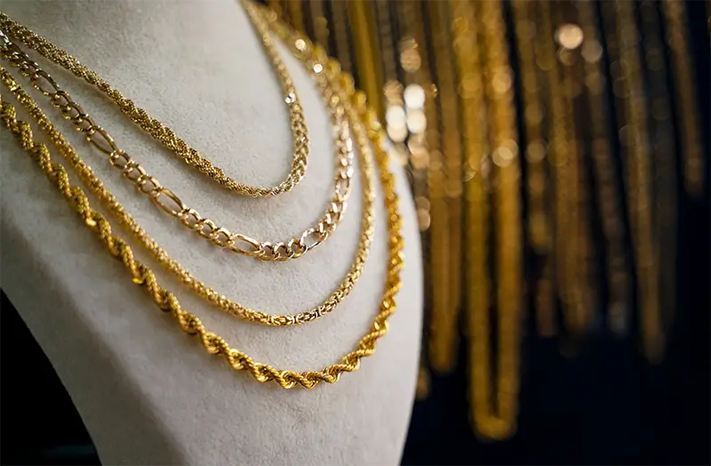 Close-up of layered gold chain necklaces in various designs, displayed on a velvet jewelry stand, with a blurred golden background.