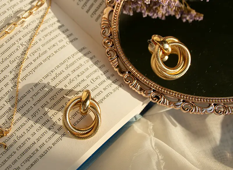 Gold jewelry, including a statement ring and chain necklace, styled on an open book with a vintage mirror, dried lavender, and warm sunlight enhancing the luxurious tones.